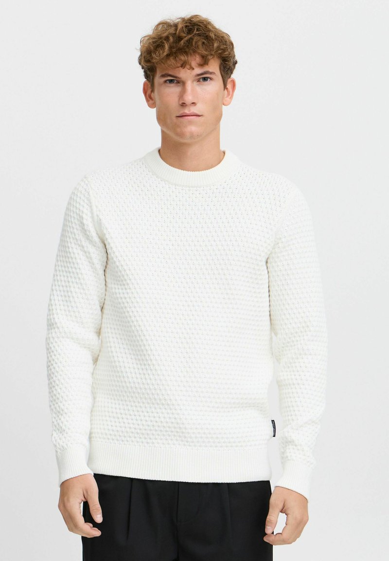 Pull en maille blanche texturée avec un col rond, des bords-côtes aux poignets et à l'ourlet, affichant un motif en nid d'abeille et une petite étiquette sur le côté.
