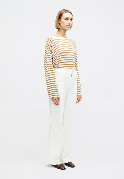 Femme debout portant un haut à manches longues à rayures beige et blanches, un pantalon blanc à jambes larges, et des chaussures plates marron sur un fond uni.