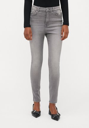 Jean slim fit gris avec une taille mi-haute, poches avant et finition délavée, assortis à des chaussures noires à talons avec des œillets décoratifs.