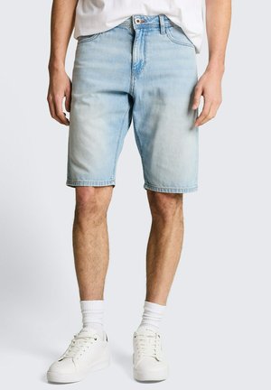 Man draagt lichtblauwe denimshorts, witte sportsokken en witte sneakers, staand tegen een effen achtergrond.
