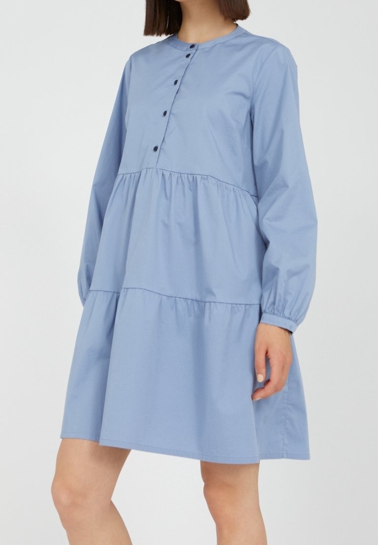 Robe en coton bleue à manches longues, col rond et boutonnage devant. Présente un design à volants avec un tissu froncé à la taille et en dessous.