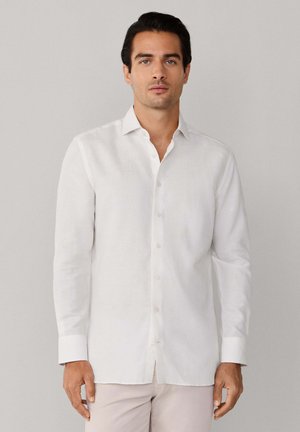 Mann trägt ein weißes Hemd mit langen Ärmeln und offenem Kragen, steht vor einem schlichten hellgrauen Hintergrund.