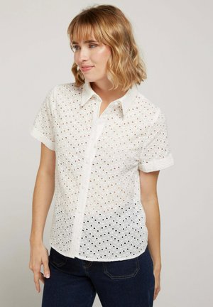 Femme portant une chemise blanche à manches courtes avec œillets et un jean bleu foncé, regardant vers sa gauche avec un léger sourire.