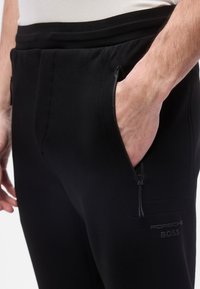 Κοντινό πλάνο ενός ατόμου που φοράει μαύρο παντελόνι φόρμας με φερμουάρ σε πλαϊνή τσέπη και λογότυπο Porsche BOSS, με το δεξί χέρι μέσα στην τσέπη.