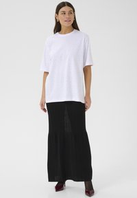 Bílé tričko s krátkým rukávem a vyšitými detaily v kombinaci s černou plisovanou maxi sukní. Outfit doplňují červené lodičky na vysokém podpatku.