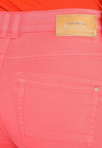 Korálově růžové džíny s kapsou vzadu a béžovou koženou etiketu s nápisem "MOS MOSH" našitou na pásku.