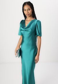 Abito da sera in satin color turchese con scollatura a ciambella e maniche a palloncino corte. Il design presenta una silhouette aderente e una texture liscia. Accompagnato da una pochette argento.