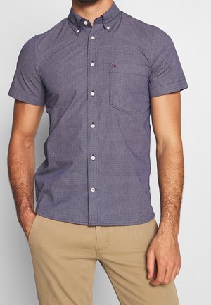 Mann trägt kurzärmeliges, marineblaues Hemd mit Muster und Brusttasche sowie beige Hose, Hände neben dem Körper ruhend.