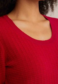 Haut tricoté rouge texturé avec un large décolleté rond. Le tissu présente un motif à carreaux subtil et des bords côtelés sur les poignets et le col.