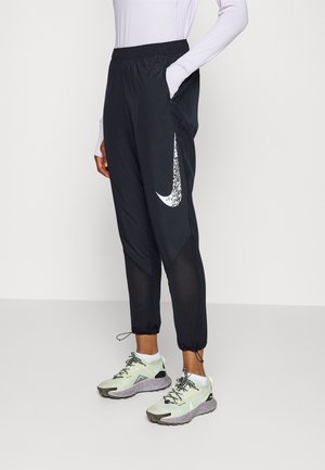 Persoon die zwarte Nike joggers draagt met een witte swoosh en meshpanelen, gecombineerd met witte sportschoenen met zwarte veters.