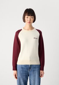 Woman with short dark hair wearing a cream sweater with burgundy sleeves and blue jeans, standing against a plain white background.