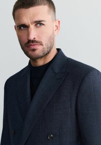 Blazer a motivo navy con finitura texturizzata, dotato di doppio bottone e rever a notched, indossato su un dolcevita nero. Ritratto in primo piano.