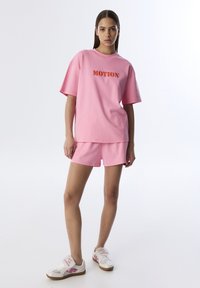 Camiseta rosa de algodón de gran tamaño con "MOTION" impreso en naranja, combinada con pantalones cortos rosa a juego. Calzado: zapatillas blancas con detalles rosa.