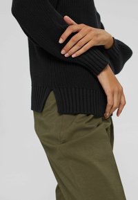 Czarny dziergany sweter z prążkowanymi mankietami i dołem, z bocznymi rozcięciami. W zestawie z oliwkowozielonymi spodniami, podkreślającymi fakturę materiału.
