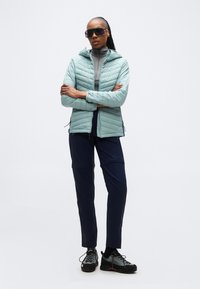 Kvinne som står med armene i kors iført en lyseblå vattert jakke, mørke bukser, tursko og store tonede solbriller på hvit bakgrunn.