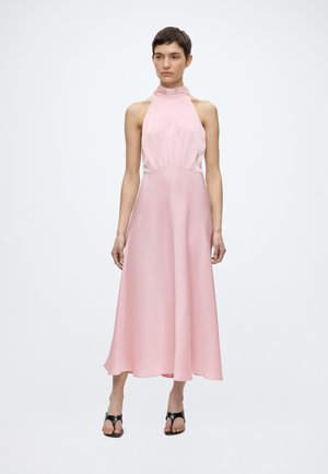 Kobieta o krótkich włosach, ubrana w różową sukienkę midi bez rękawów z wysokim kołnierzem oraz czarne czółenka z odkrytymi palcami, stojąca na jednolitym białym tle.