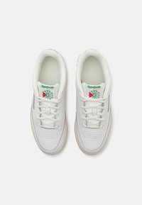 Sneaker bianche in pelle con lacci piatti, punta perforata, logo verde e rosso sulla linguetta e suola in gomma beige. Design semplice e classico.