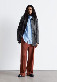 Black textured leather jacket over a light blue button-up shirt and rust-colored wide-leg trousers, paired with flat black shoes.