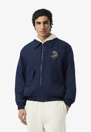 Jonge man die een marineblauwe jas met rits draagt, met een witte capuchontrui eronder en lichtgekleurde broek, kijkt recht vooruit met een neutrale uitdrukking.