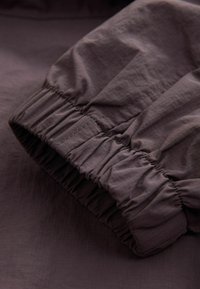 Țesătură maro cu o textură subtilă, având un manșon elastic plisat. Materialul pare ușor și neted.