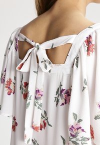 Blusa blanca con estampado floral en rosa, morado y verde. Presenta un corte cuadrado en la parte posterior y lazos en el escote, con mangas sueltas.