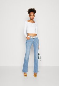 Vit långärmad croptop med en liten röd logotyp, ljusblå utsvängda jeans, vit bälte, färgglada klacksandaler och en liten iriserande väska.