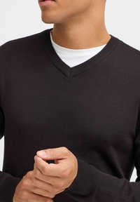 Pulover negru cu decolteu în V, realizat dintr-un material moale, cu manșete și decolteu ribbate. Modelul poartă o cămașă albă dedesubt.