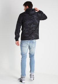 Jachetă cu glugă de camuflaj negru, dintr-un material ușor, cu mâneci lungi. Asociată cu blugi din denim bleu deschis și adidași gri sport.