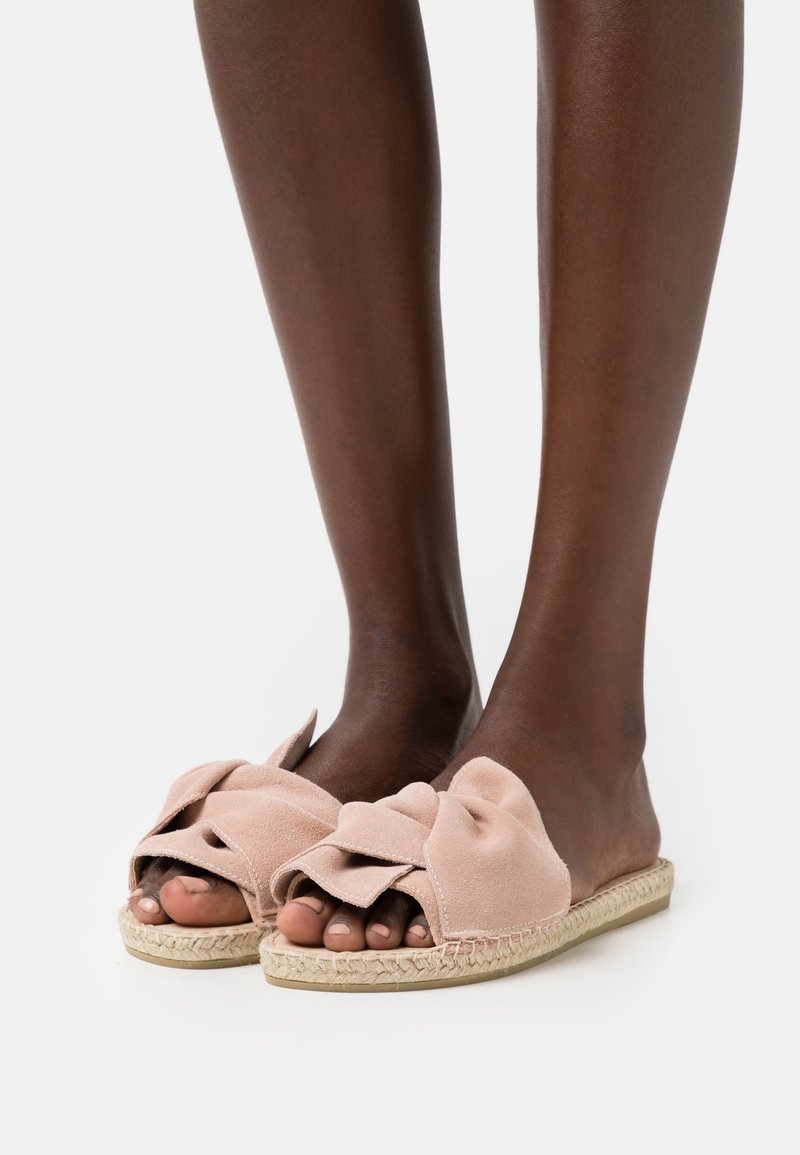 Pieds portant des sandales en daim rose pâle à enfiler avec des brides nouées et des semelles en jute tressé sur fond blanc.