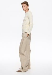 Top de manga larga en beige claro con detalles fruncidos en los laterales, combinado con pantalones holgados beige y zapatos de punta beige.