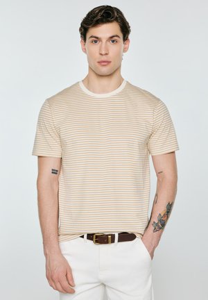 Młody mężczyzna z kręconymi włosami nosi beżowo-białą pasiastą koszulkę, białe spodnie, brązowy pasek i ma tatuaże na obu przedramionach.