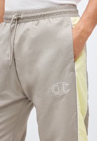 Ανοιχτό γκρι φόρμα με ελαστική μέση, κορδόνι, ανοιχτοκίτρινες πλαϊνές λωρίδες και το λογότυπο της Champion κοντά στην τσέπη, με το χέρι μέσα στην τσέπη.