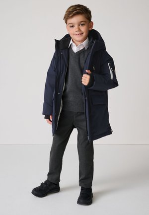 Ragazzo giovane che indossa un cappotto invernale blu navy con cappuccio sopra un maglione grigio, una camicia bianca, pantaloni grigi e scarpe nere, in piedi e sorridente.