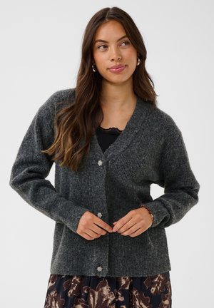 Femme portant un cardigan gris foncé boutonné sur un haut noir, une jupe à fleurs, des créoles et un bracelet, debout devant un fond clair.