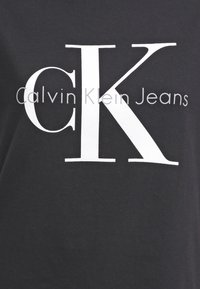 Zwarte stof met grote witte "CK" letters en kleinere grijze tekst "Calvin Klein Jeans" die gecentreerd op staat.