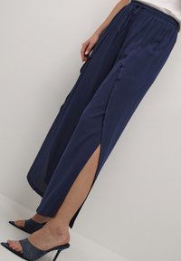 Navyblauwe wijde broek gemaakt van lichte stof, met een trekkoord in de taille en hoge zijsplitten, gecombineerd met gestructureerde hakken.