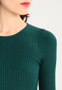 Femme portant un haut à manches longues côtelé vert avec un col rond, dévoilant son cou, des lèvres avec du rouge à lèvres rouge, et un profil partiel de son visage.