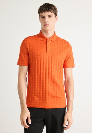 Korte mouwen polo in textuur oranje stof met een kraag en een drieknoopssluiting, gecombineerd met zwarte broek.