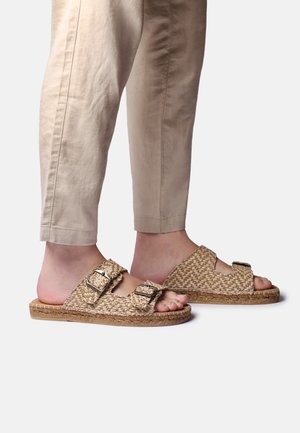 Stopy w beżowych, plecionych sandałach na płaskiej podeszwie z podwójnymi paskami z klamrami, w połączeniu z jasnokhakiowymi, krótkimi spodniami na jednolitym tle.