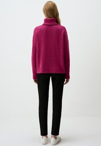 Pull en maille fuchsia oversized à col roulé avec des poignets et un ourlet côtelés ; associé à un pantalon noir slim et des baskets blanches. Fond uni.