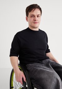 Czarna koszulka z krótkim rękawem i ciemnoszare dżinsy noszone przez osobę siedzącą na wózku inwalidzkim z zielonymi akcentami na kołach na jednolitym tle.