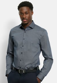 Dunkelblaues Hemd mit Knopfleiste, das ein geometrisches Muster, lange Ärmel, einen Kragen und schwarze Knöpfe aufweist. Getragen mit einem schwarzen Gürtel.