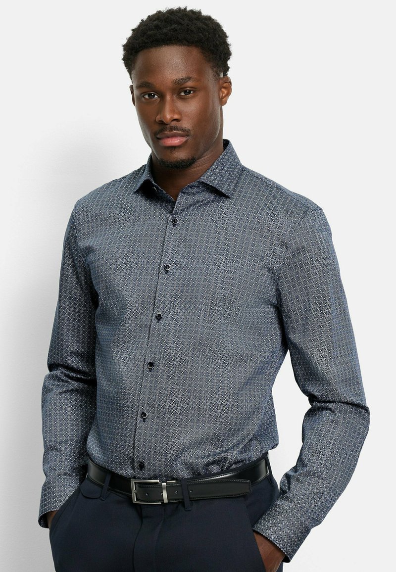 Dunkelblaues Hemd mit Knopfleiste, das ein geometrisches Muster, lange Ärmel, einen Kragen und schwarze Knöpfe aufweist. Getragen mit einem schwarzen Gürtel.