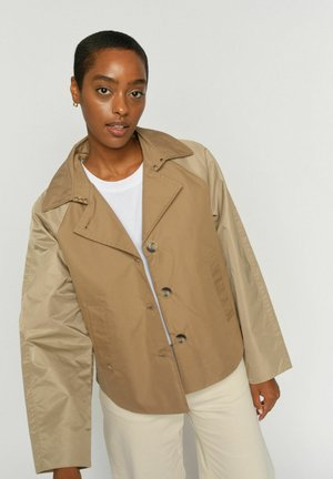 Rövid hajú fiatal, barna és bézs gombos dzsekit visel fehér ing alatt, egyszínű háttér előtt áll.