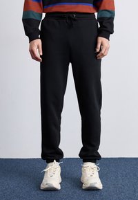 Sorte sweatpants med elastisk talje og snor. Blødt stof, tætsiddende ben, afsluttet med elastiske manchetter. Parret med lyse sneakers.