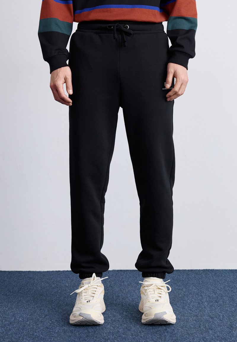 Sorte sweatpants med elastisk talje og snor. Blødt stof, tætsiddende ben, afsluttet med elastiske manchetter. Parret med lyse sneakers.