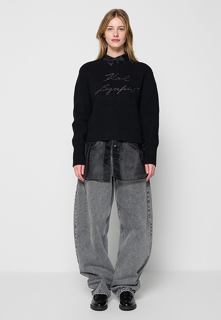 Pull en tricot noir avec texture côtelée et broderie sur le devant ; associé à un pantalon en denim gris ample et des chaussures noires, offrant un look superposé.