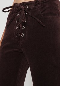 Bruna kordfläskbyxor med snörning fram och silverfärgade öglor, som framhäver en figurnära design och vertikal ribbad textur.