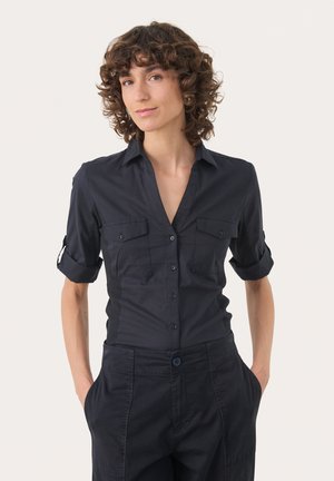 Camicia blu navy con collo a punta, maniche corte arrotolate, due tasche sul petto e una silhouette aderente. Tessuto liscio con chiusura a bottoni.