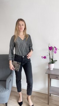 Sidabrinė blizgi ilgos rankovės palaidinė su V formos iškirpte, juodos aukštos talijos džinsai ir juodas karoliukais dekoruotas vakarėlio krepšys minimalistinėje vidinėje erdvėje.